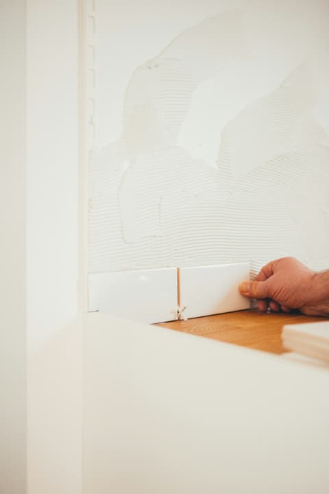 Mano colocando un azulejo blanco sobre cemento cola peinado, con crucetas para marcar la junta